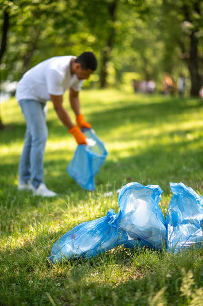 Cleaning. Young dark-skinned guy in gloves standing bent over putting waste in bag in park on sunny day - Фото, зображення