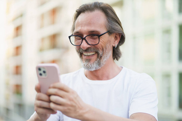 Happy caucasian middle aged man with grey beard reading or texting using smartphone standing outdoors city streets background wearing white shirt. Middle aged man on street of european city. - Photo, image
