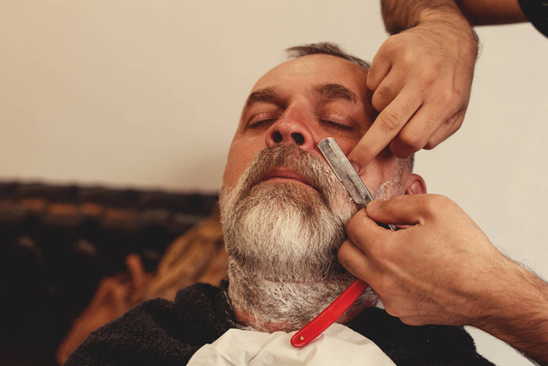 Shaving with a straight razor in a barbershop. A bearded old man being shaved in a barbershop. Classic shave by Stainless Steel Straight Edge Razor. - Foto, immagini