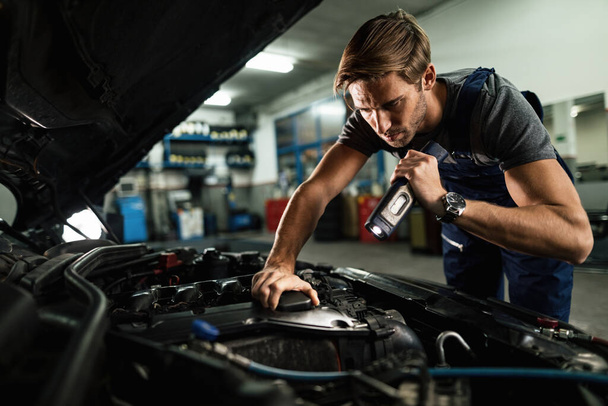 Young mechanic using lamp while examining malfunction of car engine in auto repair shop.  - Photo, Image