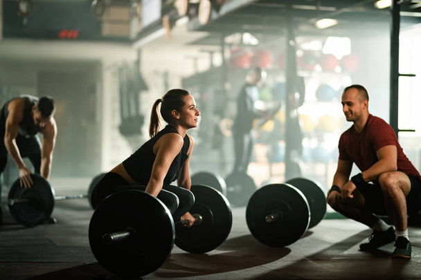 Young athletic woman lifting barbell while having strength training with her instructor in a gym.  - Foto, afbeelding