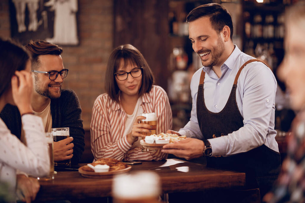Молодой счастливый официант угощает клиентов, которые пьют пиво в пабе.  - Фото, изображение