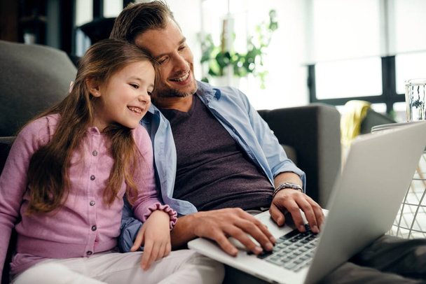 Happy father and daughter sitting close to each other while using computer and surfing the internet at home.  - 写真・画像