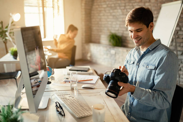 Young happy photographer looking at photos on his camera while working in the office.  - 写真・画像