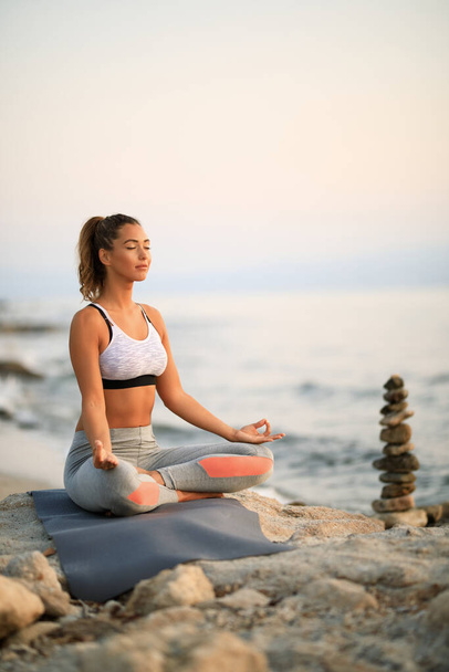 Young athletic woman practicing Yoga with eyes closed while sitting in lotus position on rocky beach.  - 写真・画像