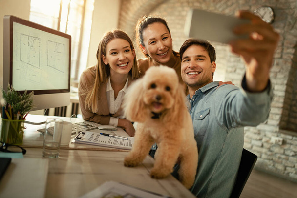 Team of freelance workers having fun while taking selfie with a poodle in the office.  - Zdjęcie, obraz