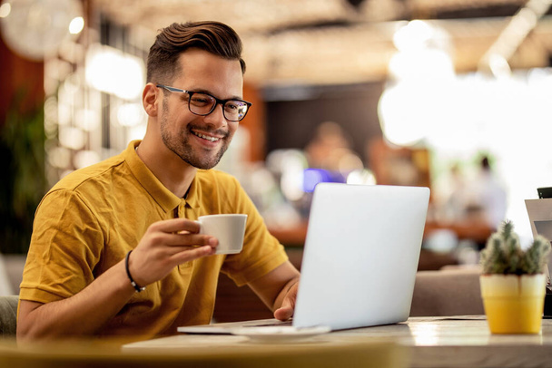 Smiling man surfing the net on laptop while drinking coffee in a bar.  - Zdjęcie, obraz