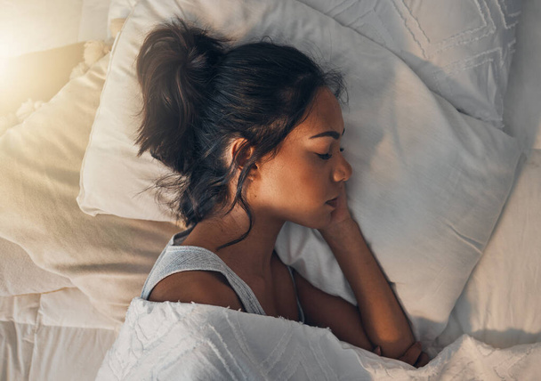 Красивая молодая смешанная расовая женщина спит в мягкой удобной кровати дома. Одна испаноязычная женщина чувствует себя измученной и отдыхающей в успокаивающей постели. - Фото, изображение