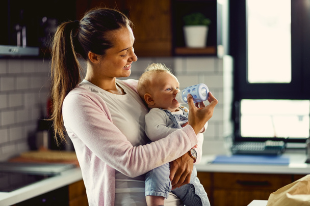 Молодая заботливая мать использует детскую бутылочку и кормит своего маленького сына на кухне.  - Фото, изображение