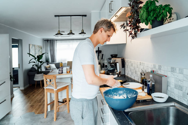 A young man is cooking cheese pancakes in a modern light kitchen at home. Man preparing pancakes for frying on the pan. Housework and home duties. Culinary hobby. Selective focus - Foto, imagen