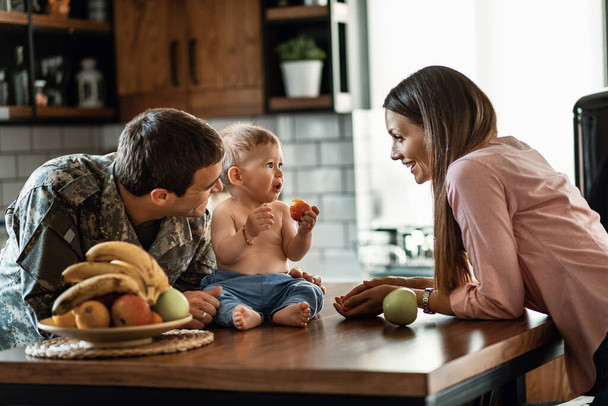 Happy military officer and his wife spending time with their baby son who is eating fruit at home.  - Fotoğraf, Görsel
