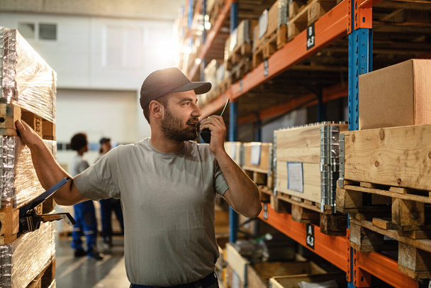 Male warehouse worker communicating with someone over walkie-talkie while standing among shelfs in distribution warehouse.  - Photo, image