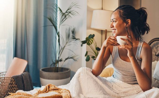 Una hermosa joven hispana disfrutando de una cálida taza de café para desayunar. Una hembra de raza mixta tomando té mientras está sentada en la cama y soñando despierto. - Foto, Imagen