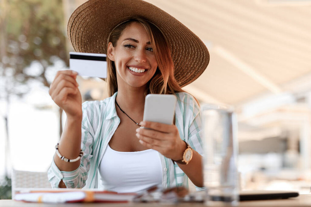 Happy woman with credit card checking her online back account on smart phone while relaxing in a cafe.  - Photo, Image