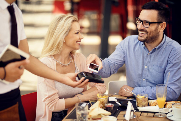 Щасливий чоловік розміщує смартфон на зчитувачі кредитних карток і робить безконтактну оплату в барі
.  - Фото, зображення