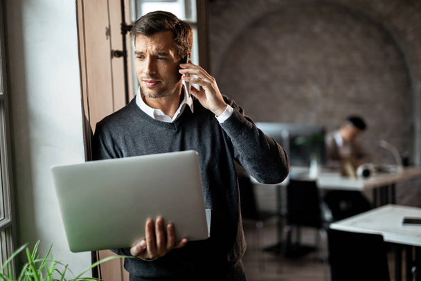 Бизнесмен держит ноутбук и разговаривает по мобильному телефону, стоя у окна в офисе.  - Фото, изображение