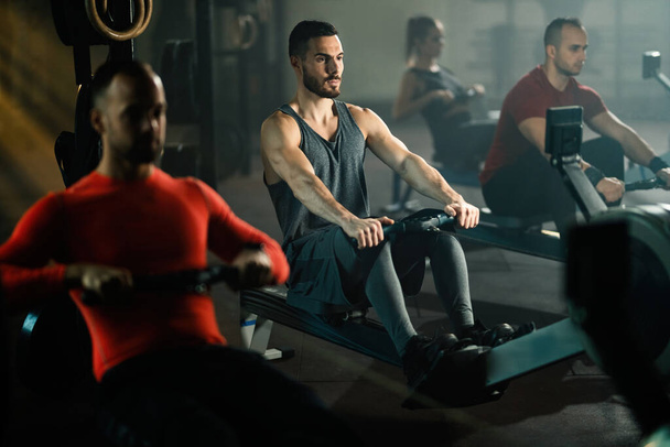 Small group of athletic people having sports training on rowing machines in a gym. Focus is on man in the middle.  - Zdjęcie, obraz