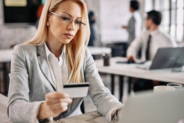Σοβαρή επιχειρηματίας πιστωτική κάρτα και φορητό υπολογιστή για να ψωνίσετε online, ενώ κάθεται στο γραφείο της. - Φωτογραφία, εικόνα