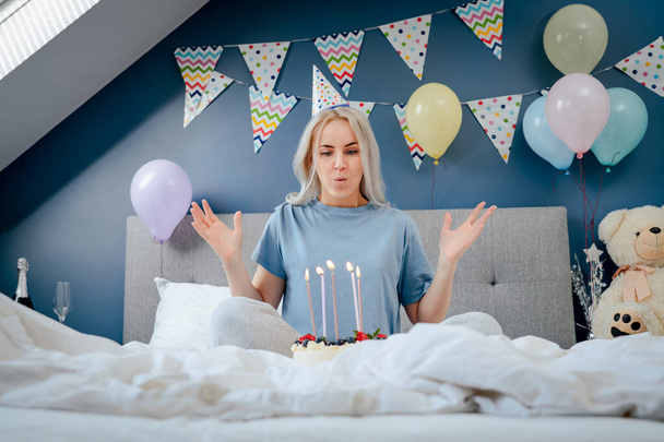 Glückliche Frau in Pyjama und Partymütze, die Kerzen auf der Geburtstagstorte auf dem Bett im geschmückten Schlafzimmer ausbläst. Machen Sie einen Wunschprozess. Überraschung am Morgen. Happy Birthday Konzept. Selektiver Fokus - Foto, Bild