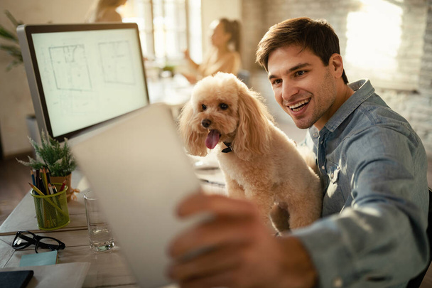 Щасливий позаштатний працівник розважається, використовуючи тачпад і беручи селфі зі своїм собакою в офісі
.  - Фото, зображення