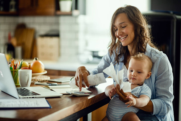 Young working mother with baby using calculator and going through bills while calculating home finances.  - Foto, Bild
