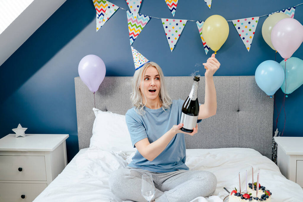Happy emotional woman in pajama and party cap opening bottle of champagne, sitting on the bed with festive cake in a decorated bedroom. Time for yourself. Celebration at home. Happy birthday concept - Zdjęcie, obraz