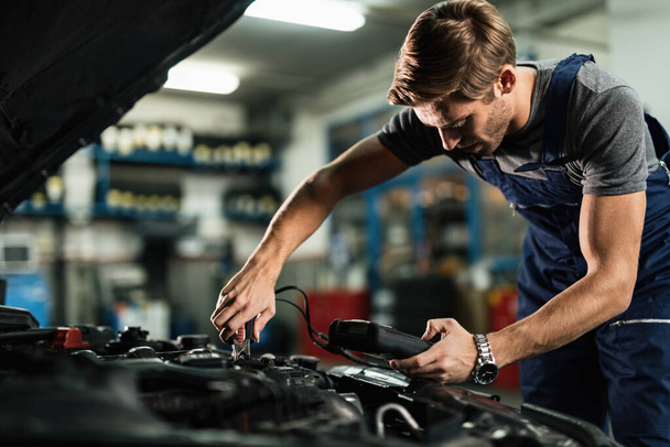 Young mechanic examining car battery with diagnostic tool while working in auto repair shop.  - Foto, Bild