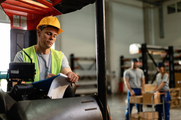 Mid adult forklift operator writing shipment reports while working in distribution warehouse.  - Valokuva, kuva