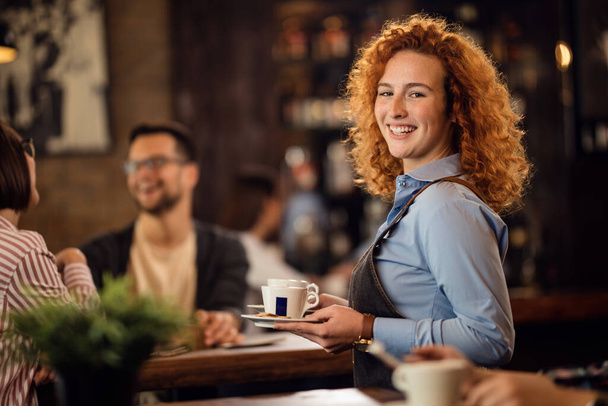 Щаслива руда офіціантка працює в кафе і подає гостям каву, дивлячись на камеру
.  - Фото, зображення