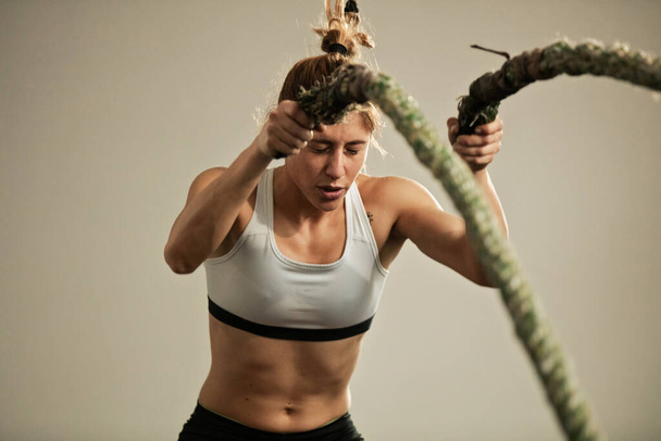 Dedicated female athlete having cross training in gym and exercising with battle rope. - Foto, immagini