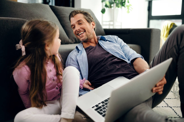 Happy father using laptop and having fun while being tickled by his daughter at home.   - Fotoğraf, Görsel