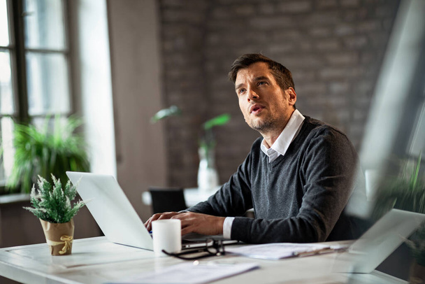Предприимчивый бизнесмен сидит на ноутбуке и смотрит вверх, думая о чем-то в офисе.  - Фото, изображение