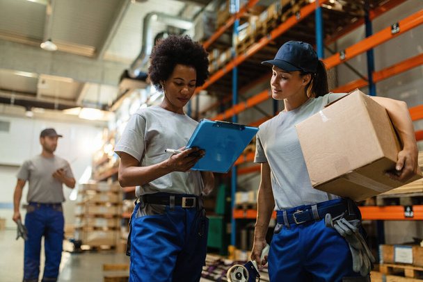 Low angle view of two female workers reading delivery schedule list while working in distribution warehouse.  - Foto, imagen