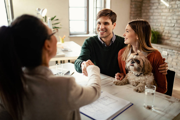 Happy couple with a dog shaking hands with real estate agent in the office. - Photo, image
