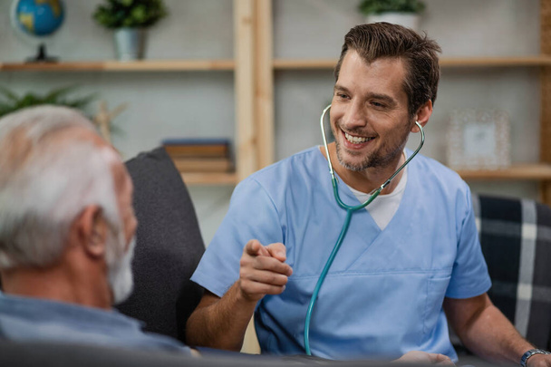 Happy healthcare worker communicating with senior man and pointing at him while being in a home visit.  - Foto, Imagen