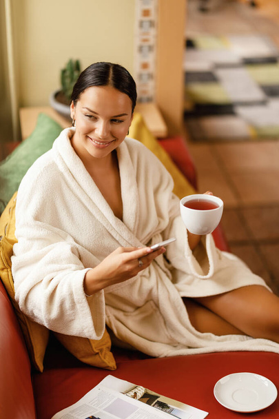 Молодая улыбающаяся женщина в халате пьет чай и пользуется смартфоном во время отдыха в оздоровительном центре.  - Фото, изображение
