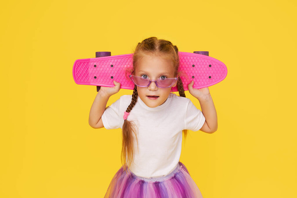 A cute little girl 6 years old in a bright tulle skirt and a white t-shirt and stylish glasses holds a bright colored skateboard on a yellow background - 写真・画像