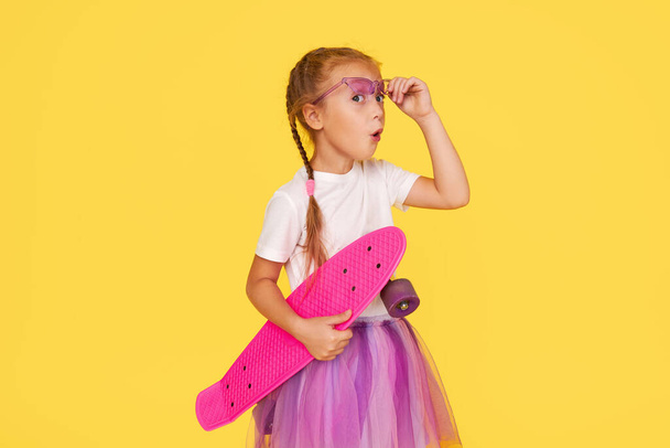 A cute little girl 6 years old in a bright tulle skirt and a white t-shirt and stylish glasses holds a bright colored skateboard on a yellow background - Foto, Imagen