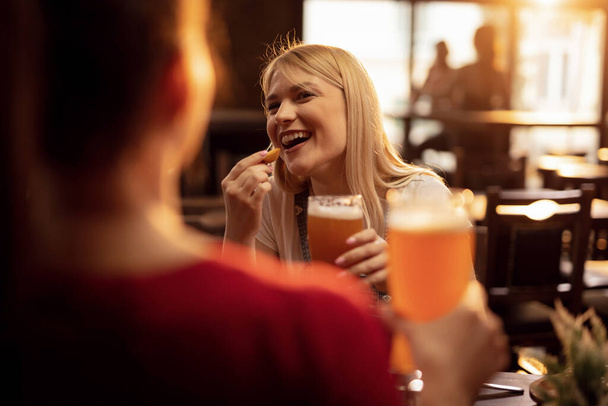 Счастливая женщина веселится во время еды и питья пива со своей подругой в таверне.  - Фото, изображение