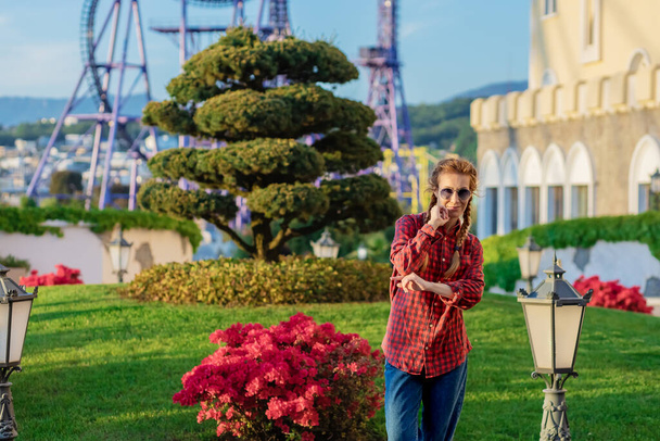 Nuorisopaidassa ja farkuissa oleva nainen seisoo kukkapenkin vieressä huvipuiston taustaa vasten. - Valokuva, kuva