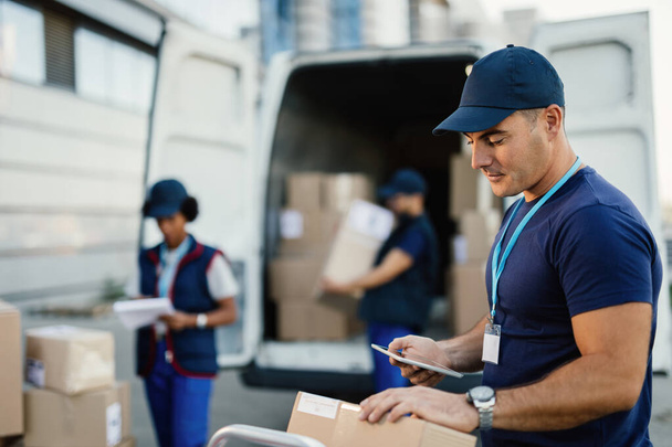 Young delivery man checking package while using digital tablet outdoors. His coworkers are unloading boxes from a van in the background.  - Foto, imagen