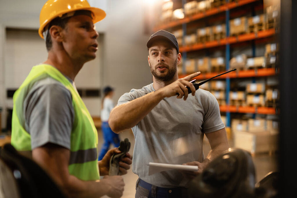 Young dispatcher communicating about delivery schedule with warehouse worker in factory warehouse.  - Photo, Image