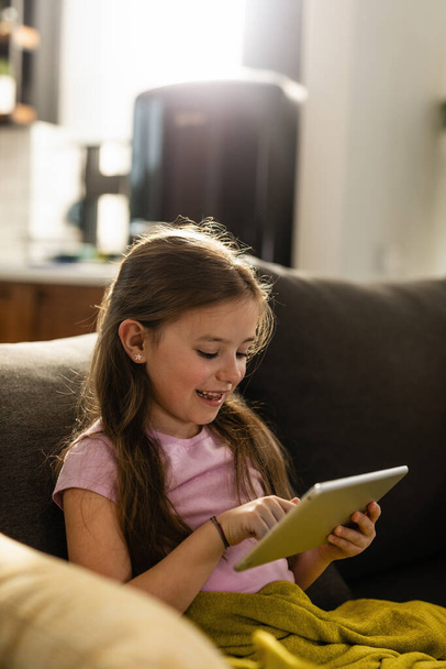 Happy little girl using touchpad and playing games while relaxing on the sofa at home.  - Foto, afbeelding