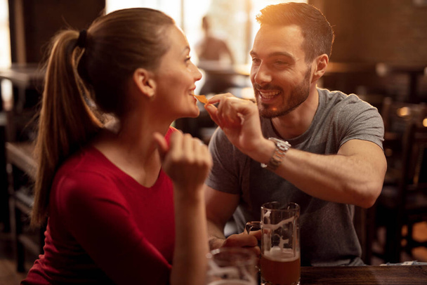 Happy man feeding his girlfriend with nacho chips while drinking beer together in a pub.  - Foto, imagen