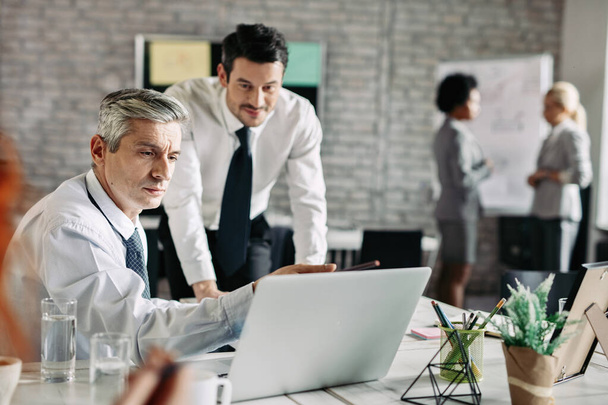 Средний взрослый бизнесмен и его младший коллега сотрудничают во время использования ноутбука в офисе.  - Фото, изображение