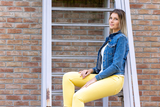 Стильная брюнетка в джинсовой жилетке, белой футболке и желтых джинсах, позирующая на фоне уличной, городской одежды. Уличная фотография. Высокое качество фото - Фото, изображение