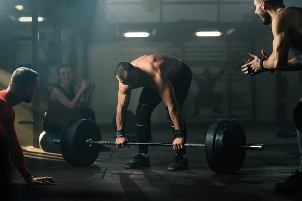 Group of athletes supporting muscular build man who is performing deadlift with barbell in a gym.  - 写真・画像