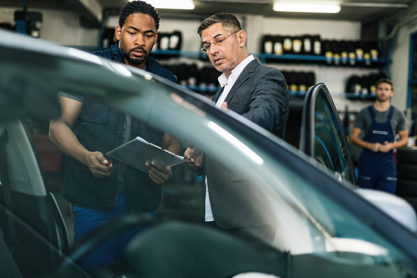 Молодой черный автомеханик и его клиент общаются во время осмотра автомобиля в мастерской.  - Фото, изображение