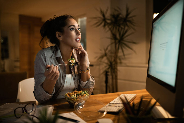 Молода жінка їсть салат під час серфінгу в Інтернеті на комп'ютері ввечері вдома
.  - Фото, зображення