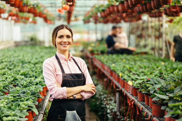 Happy female florist standing with arms crossed and looking at camera at plant nursery. There are people in the background.  - Zdjęcie, obraz